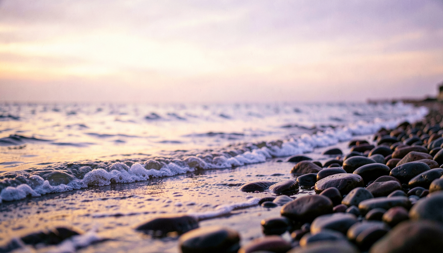 A quiet shoreline at dawn with gentle waves lapping against smooth stones, soft lavender and pale gold light spreading across the horizon, calm and hopeful