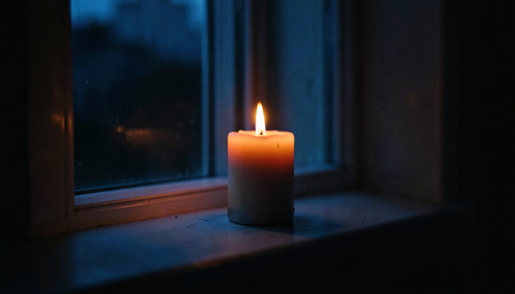 A softly lit candle on a quiet windowsill at dusk, warm amber glow against deep blue shadows, calm and contemplative mood